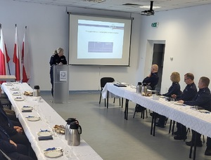 Odprawa roczna w Sępólnie Krajeńskim. Policjanci oraz zaproszeni goście siedzą przy stołach. Przy mównicy stoi Komendant Powiatowy policji w Sępólnie Krajeńskim młodszy inspektor Klaudia Ryczek. W tle wyświetlana jest prezentacja multimedialna.