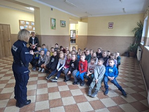 Policjantka stoi przed uczniami siedzącymi na ławeczkach. Zwrócona jest do zdjęcia plecami.
