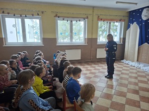 Policjantka stoi przed uczniami siedzącymi na ławeczkach. Zwrócona jest do zdjęcia bokiem.
