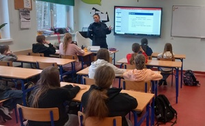 Policjantka prowadzi lekcje  w klasie lekcyjnej. Dzieci siedzą w ławkach szkolnych a policjantka stoi przed nimi. Za jej plecami wyświetlana jest prezentacja multimedialna.