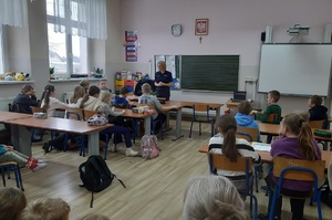 Sala lekcyjna. Policjantka stoi przed uczniami siedzącymi w ławkach szkolnych. Dzieci zwrócone są do zdjęcia plecami.