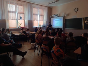 Sala lekcyjna. Policjantka stoi przed uczniami siedzącymi w ławkach szkolnych. Dzieci zwrócone są do zdjęcia plecami.