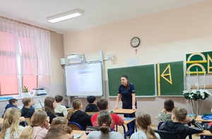 Sala lekcyjna. Policjantka stoi przed uczniami siedzącymi w ławkach szkolnych. Dzieci zwrócone są do zdjęcia plecami.