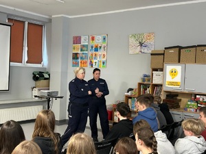 Policjantki stoją na środku sali lekcyjnej Szkoły Podstawowej nr 1 w Więcborku. Za nimi na ścianie wyświetlana jest prezentacja multimedialna. Przed policjantkami, na ustawionych w rzędach krzesłach, siedzą uczniowie. Funkcjonariuszka zwrócona jest przodem do zdjęcia.
