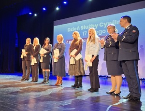 Scena sali kinowej w Centrum Kultury i Sztuki w Sępólnie Krajeńskim. Wspólne zdjęcie policjantów z wyróżnionymi pracownikami z okazji Obchodów Dnia Służby Cywilnej. W tle wyświetlany  jest slajd Dzień Służby Cywilnej 17 luty 2026 rok.