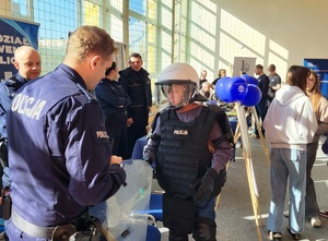Drzwi otwarte w liceum ogólnokształcącym w Sępólnie Krajeńskim. Uczniowie stoją przy policyjnych stoiskach na których ułożony jest  sprzęt wykorzystywany w służbie. Cześć z uczniów rozmawia z policjantami. W tle widać ustawione policyjne rolapy. Jeden z uczni ubiera sprzęt przeciwuderzeniowy.