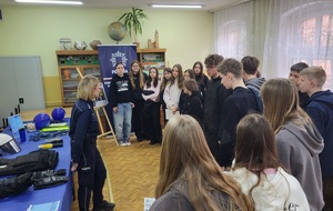 Policjantka prowadzi spotkanie z uczniami dotyczące promowania zawodu policjanta. W tle widać stoisko oraz banery policyjne.