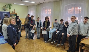 Policjantka prowadzi spotkanie z uczniami dotyczące promowania zawodu policjanta. W tle widać stoisko oraz banery policyjne.