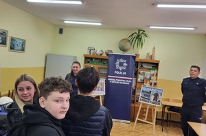 Policjantka prowadzi spotkanie z uczniami dotyczące promowania zawodu policjanta. W tle widać stoisko oraz banery policyjne.