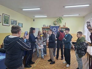 Policjantka prowadzi spotkanie z uczniami dotyczące promowania zawodu policjanta. W tle widać stoisko oraz banery policyjne.
