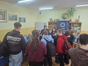 Policjantka prowadzi spotkanie z uczniami dotyczące promowania zawodu policjanta. W tle widać stoisko oraz banery policyjne.