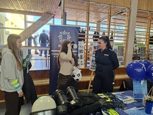 Policjantka stoi przy stoisku razem z innymi uczestnikami drzwi otwartych. W tle widać baner policyjny.